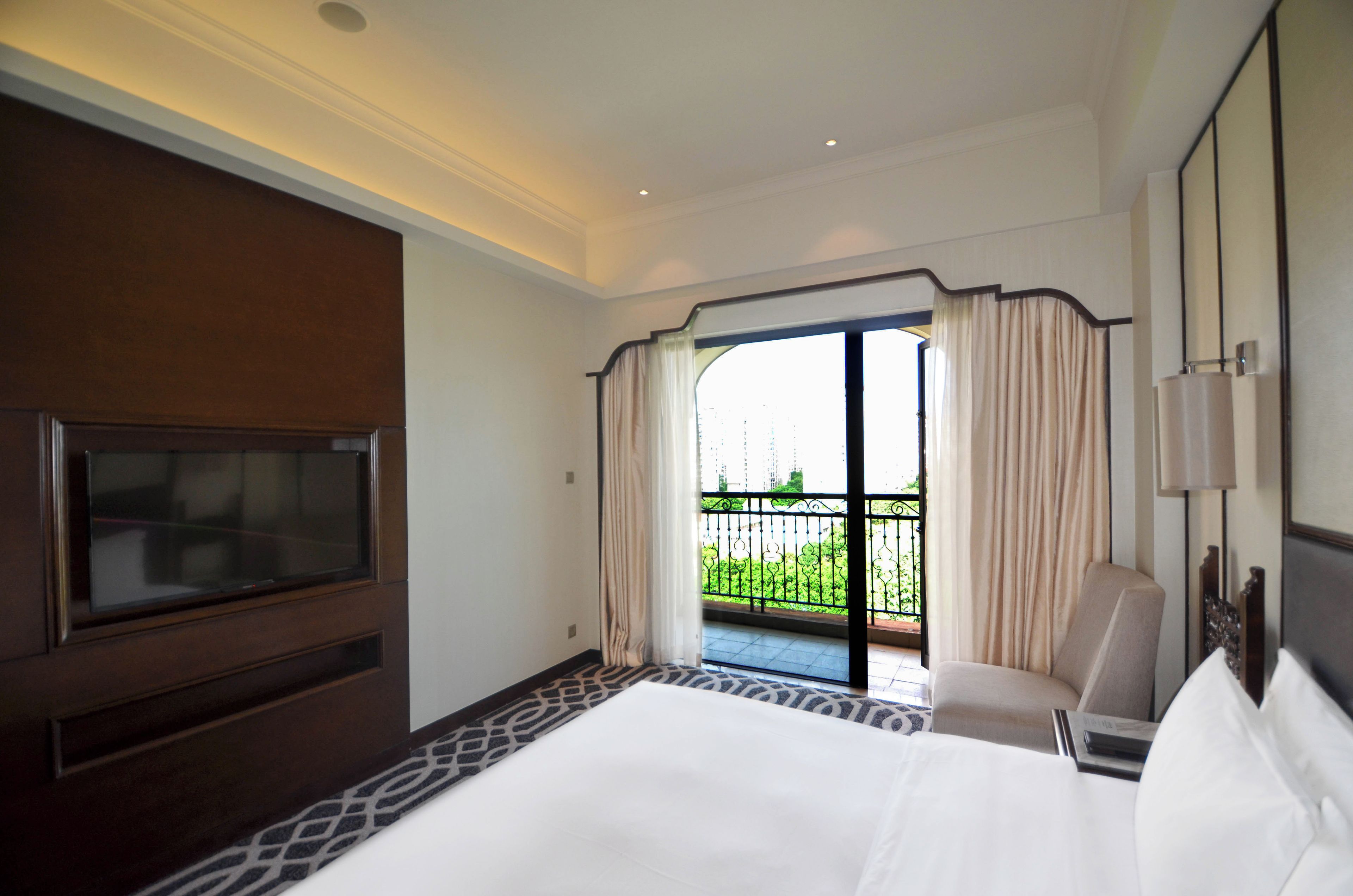 Habitación de hotel con una cama grande, televisión, una silla, un balcón con cortinas y vistas a zonas verdes.