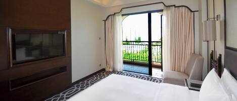 Habitación de hotel con una cama grande, televisión, una silla, un balcón con cortinas y vistas a zonas verdes.