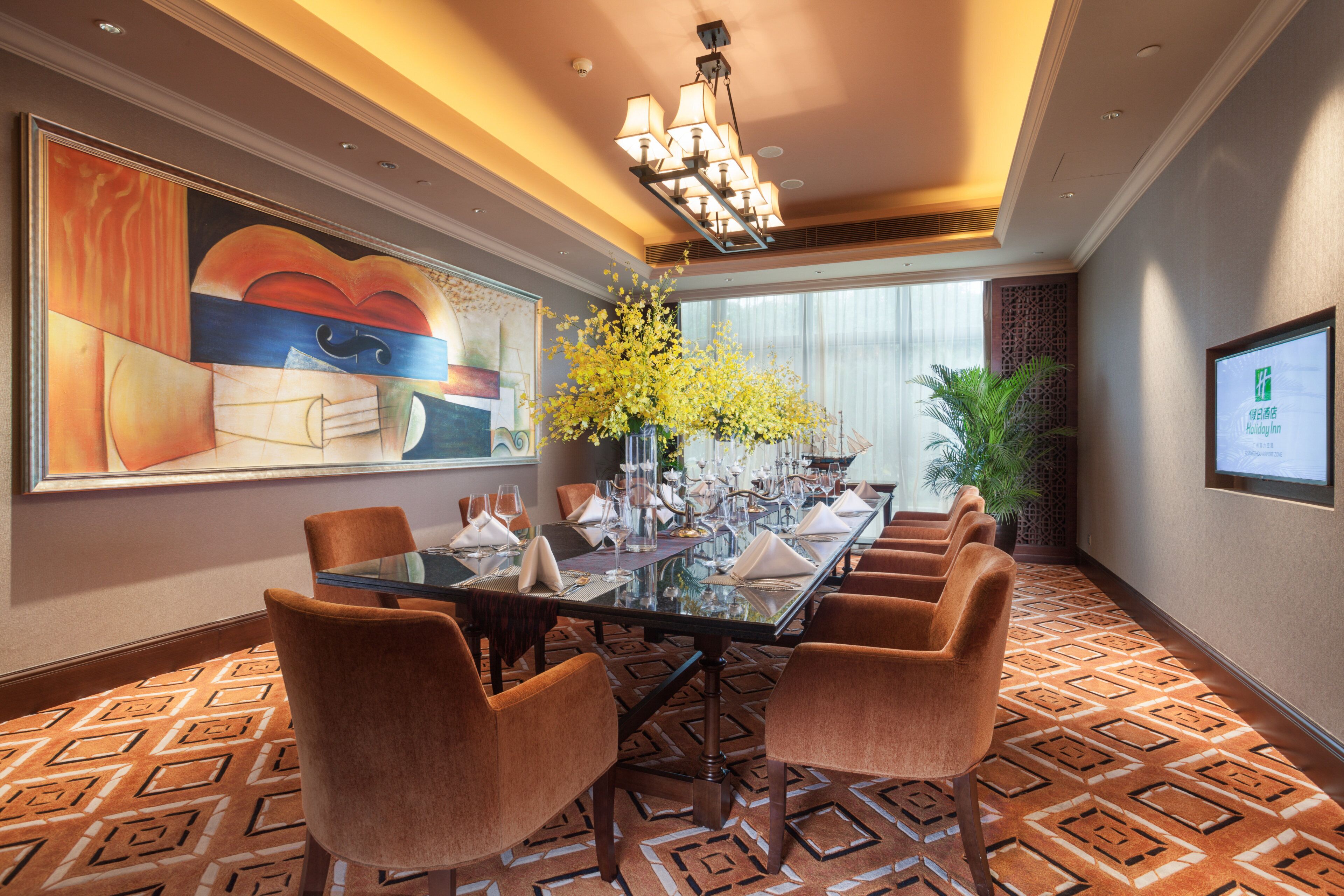 A conference room with a glass table, brown chairs, a large abstract painting, a yellow flower arrangement, and a Holiday Inn sign.