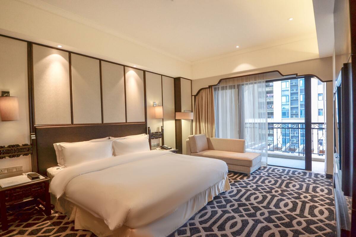 A hotel room with a large bed, a sofa, a desk, and a window with a view of buildings.