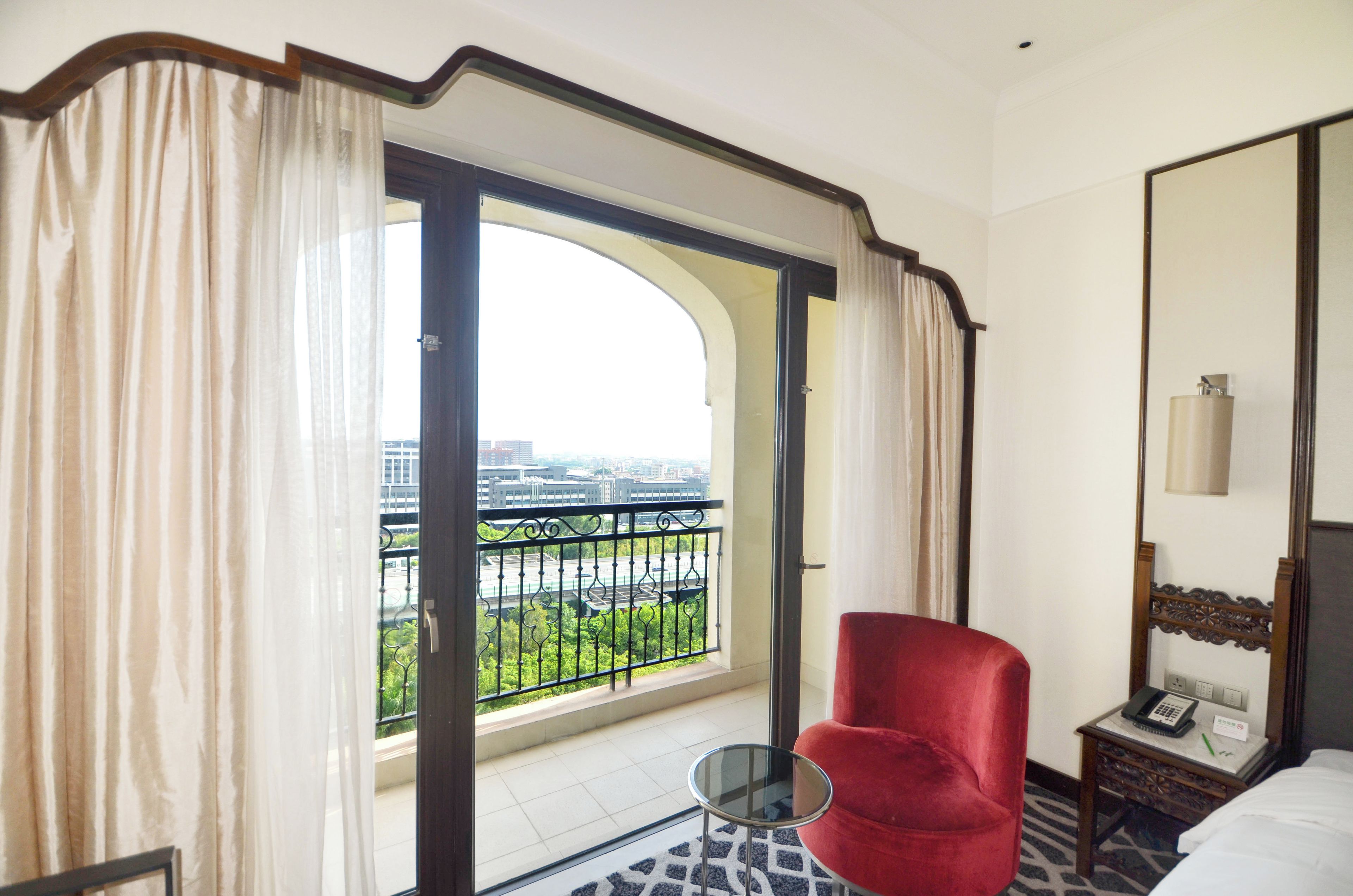 A hotel room with a large window, a red chair, a small table, and a bed.