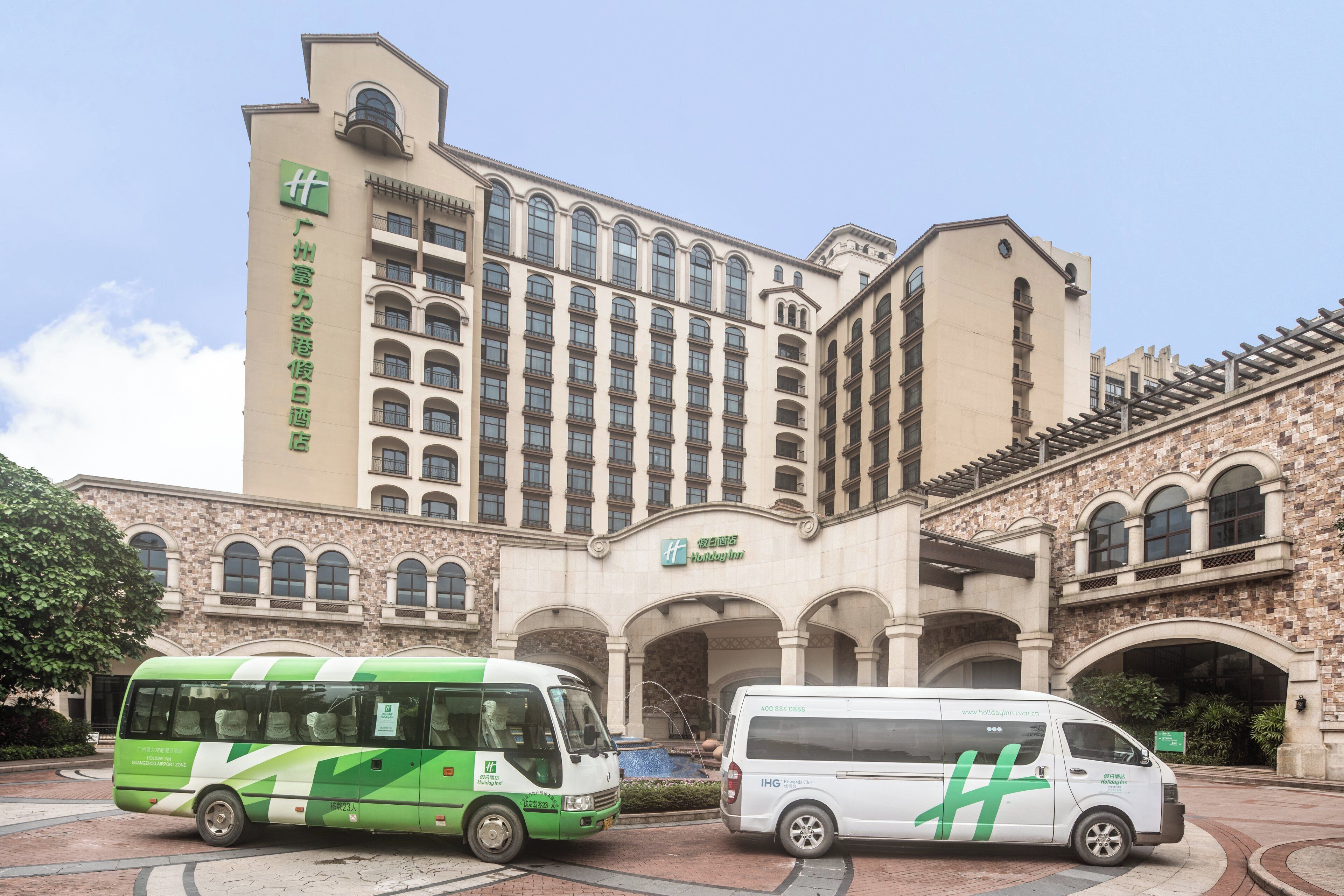 Un edificio con un cartel grande de Holiday Inn, un autobús verde y blanco, y una camioneta blanca estacionados al frente.