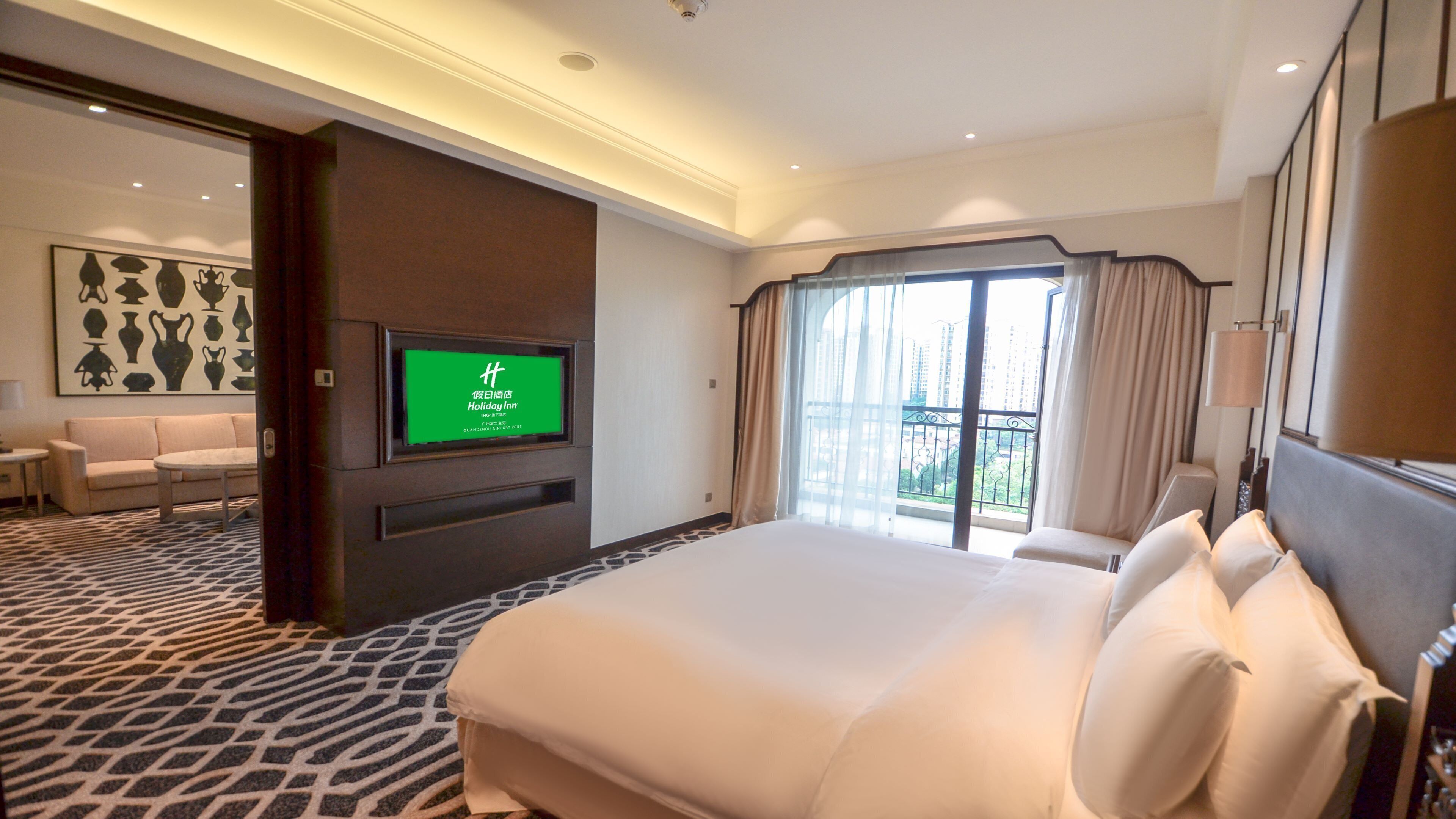 Habitación de hotel con una cama grande, un televisor montado en la pared, un balcón con cortinas y una alfombra estampada.
