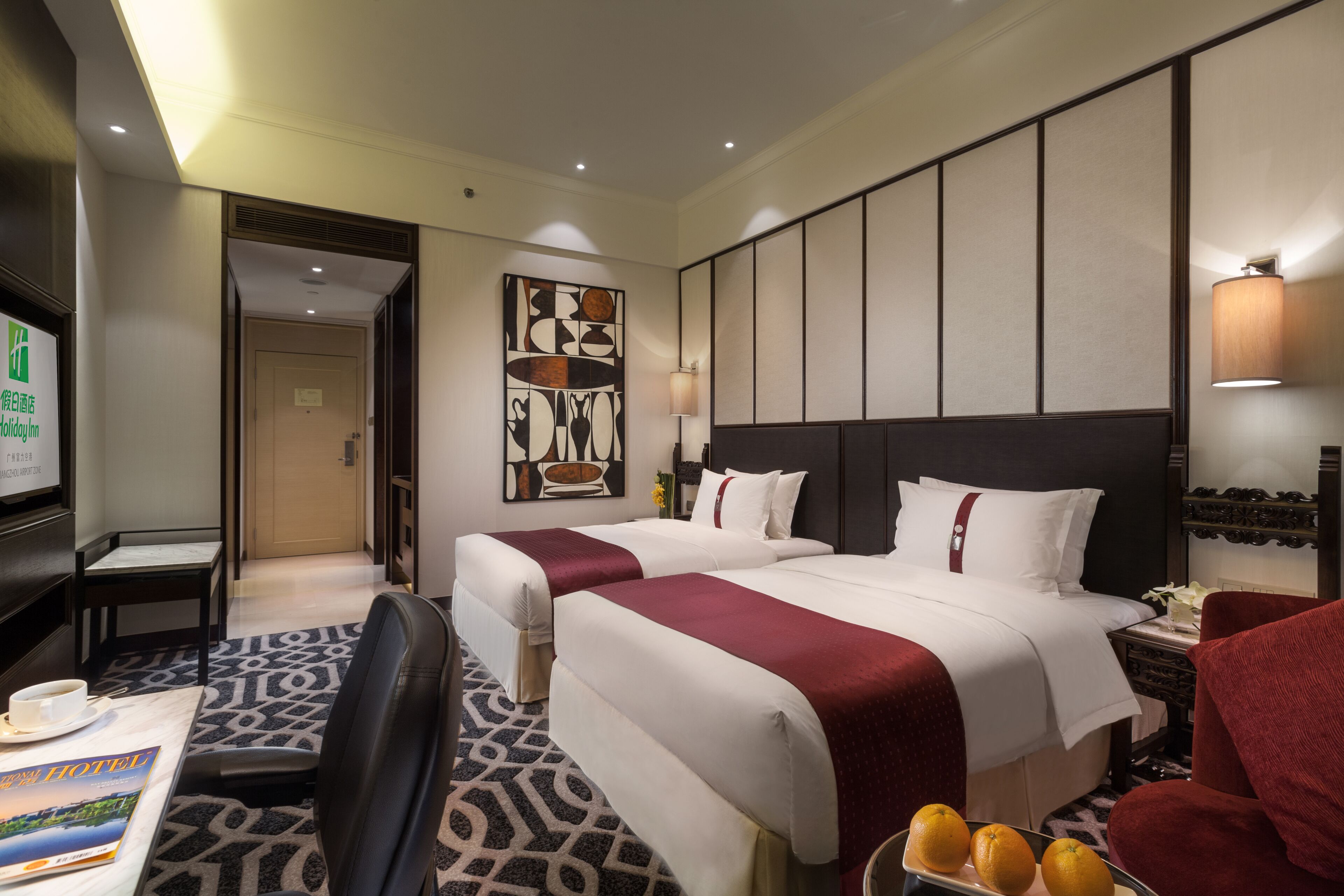 A hotel room with two beds, a desk, a chair, a TV, and a wall-mounted lamp.