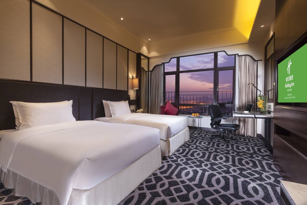 A hotel room with two beds, a desk with a chair, a large window with a view, and a television on the wall.