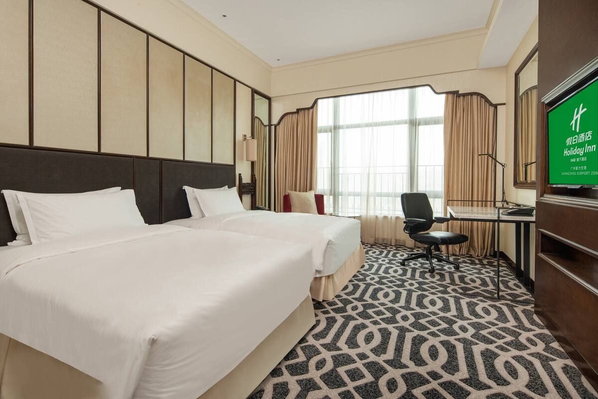 A hotel room with two beds, a desk, a chair, a TV, and a large window with curtains.