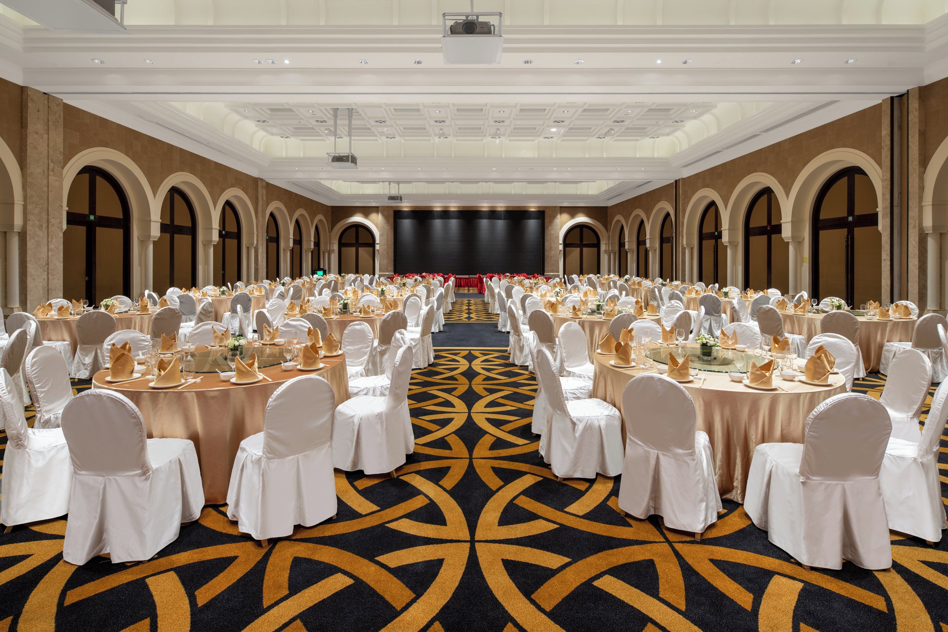 Un salón de banquetes con mesas redondas, sillas blancas y una alfombra estampada.