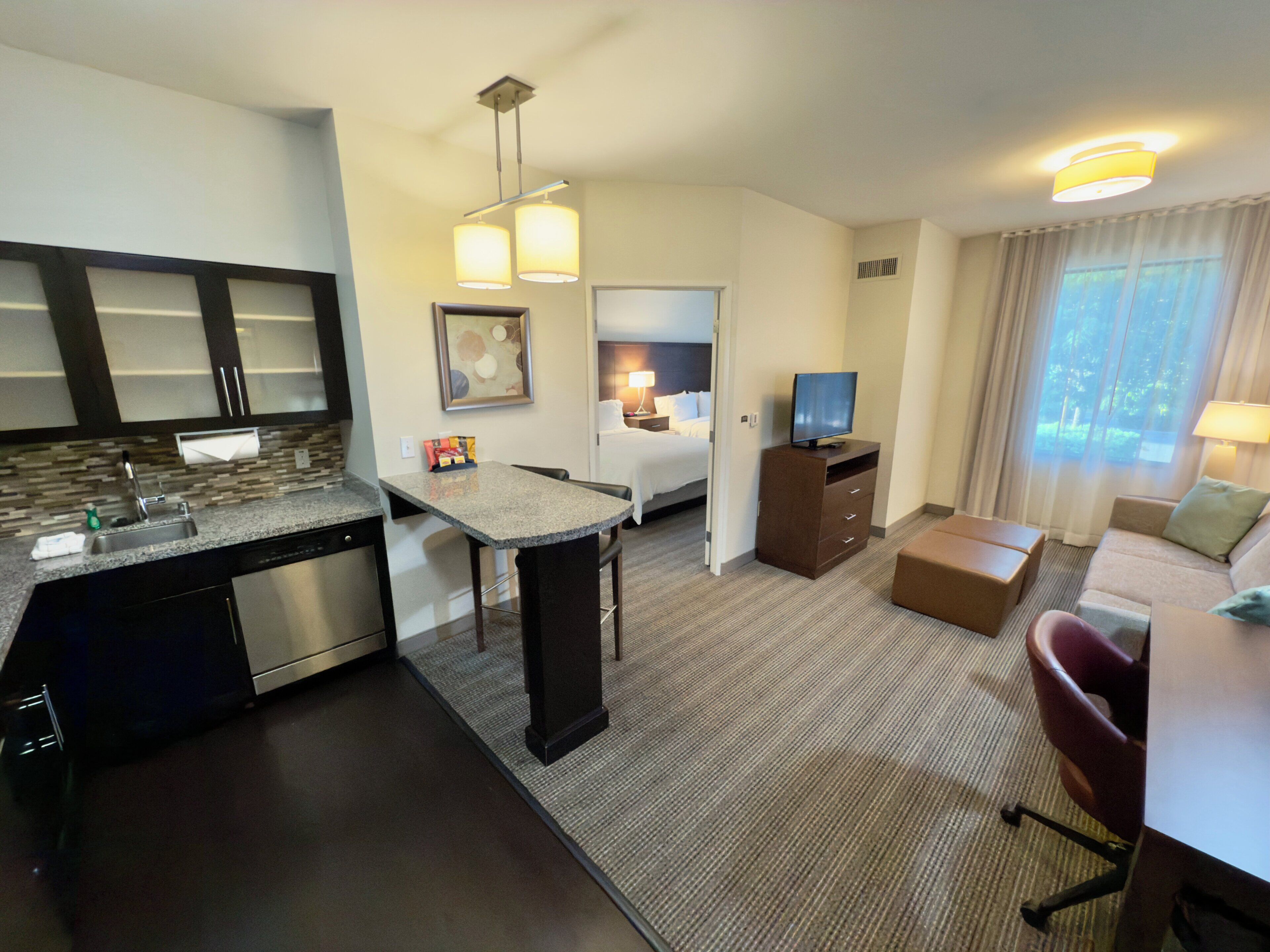 A hotel room with a kitchenette, a dining area, a bed, a TV, and a seating area with a view of the outdoors.