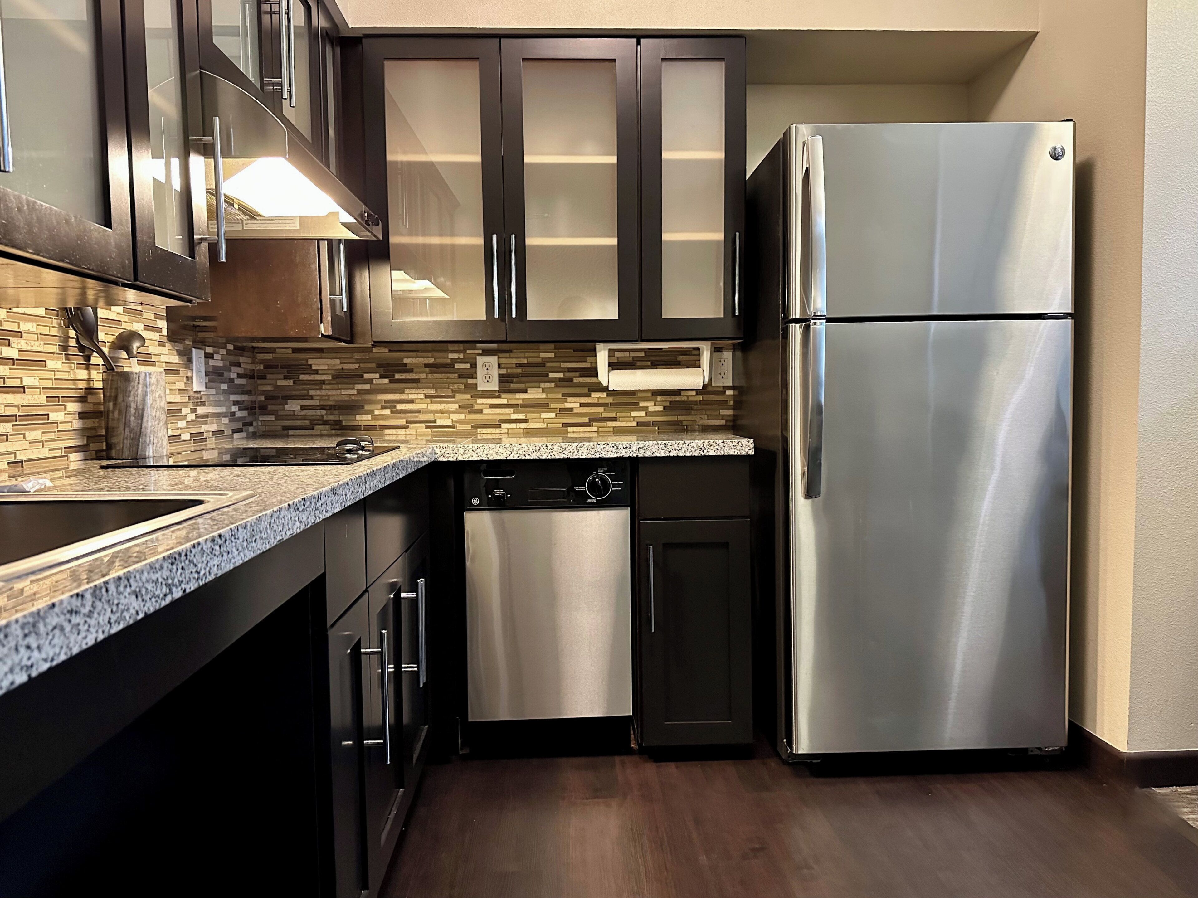 A modern kitchen with dark cabinets, a stainless steel refrigerator, a dishwasher, and a granite countertop.