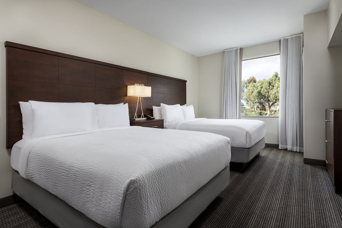 Habitación de hotel con dos camas, una mesita de noche con lámpara, cómoda y un amplio ventanal con cortinas.