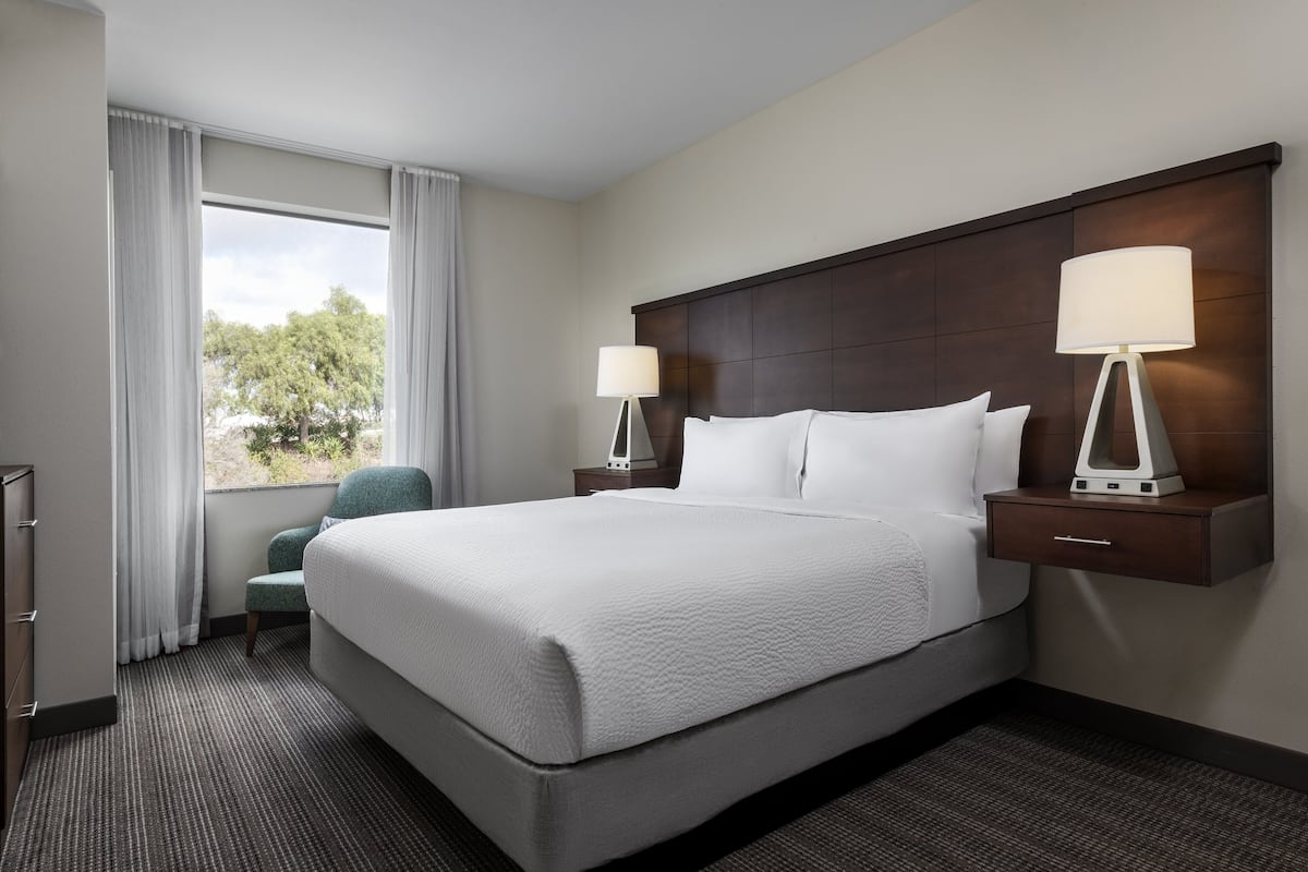 Habitación de hotel con una cama grande, dos mesitas de noche con lámparas, una silla, un aparador y una ventana con cortinas.
