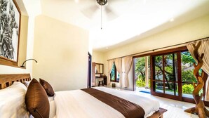 A spacious bedroom with a large bed, a wooden bench, a desk, and a view of a lush garden through large windows. - Ocean Sun Dive Resort (Tulamben)