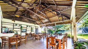 A spacious outdoor café with wooden tables and chairs, a bar area, and a view of a garden. - Ocean Sun Dive Resort (Tulamben)