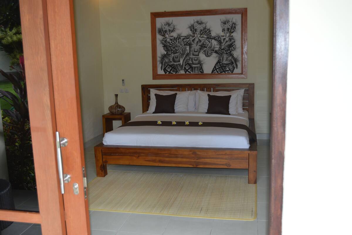 A wooden bed with white and dark brown bedding, a framed artwork above it, and a woven mat on the floor.