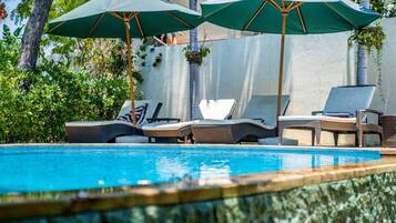 A poolside area with lounge chairs and umbrellas, surrounded by greenery and a white wall.