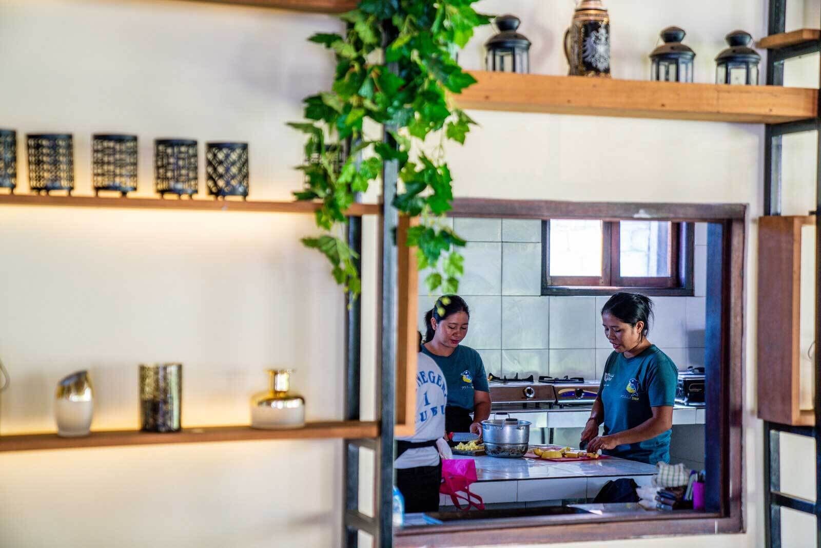 A kitchen with two people cooking, shelves with decorative items, and a window with greenery.