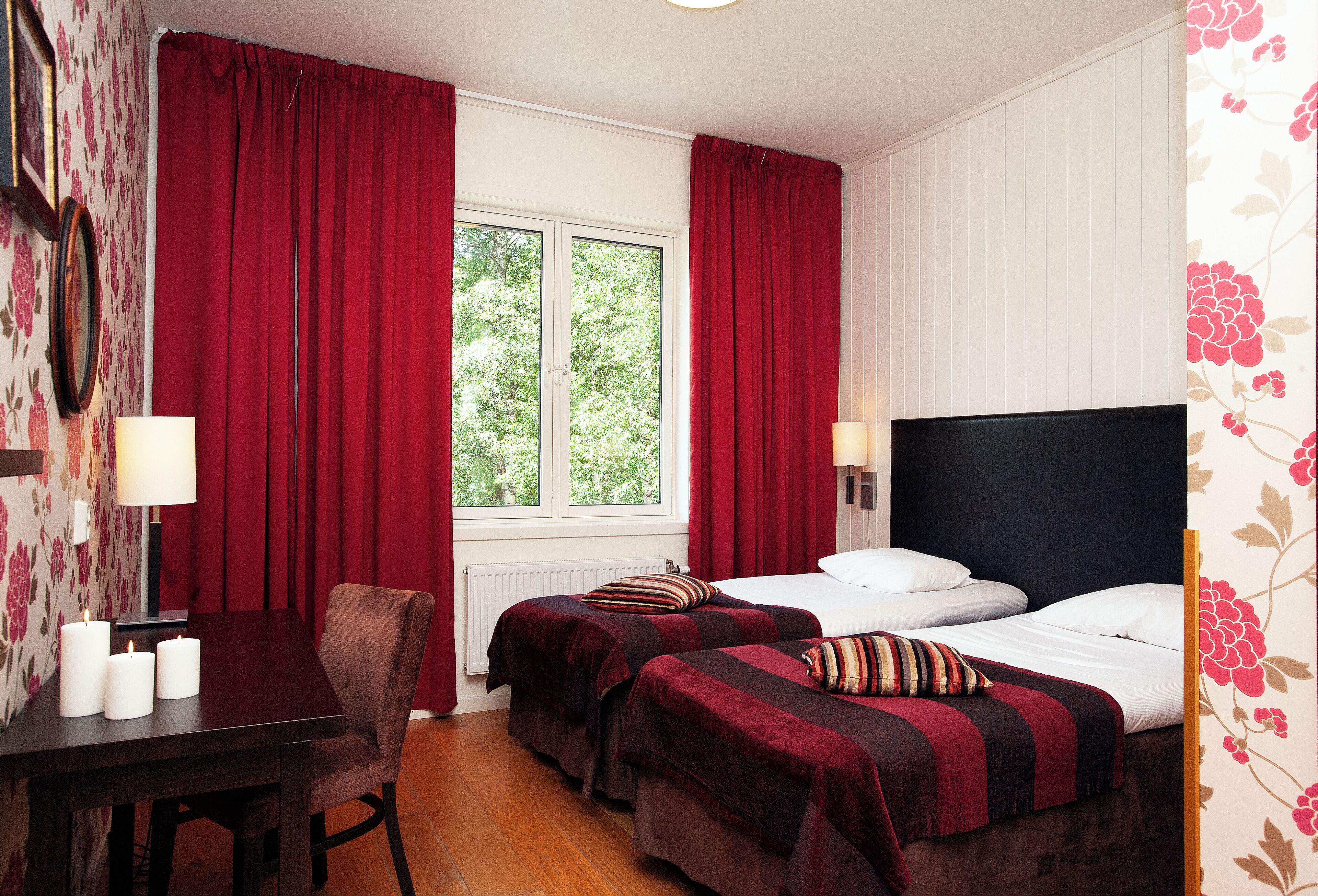 Habitación de hotel con dos camas, un escritorio con silla, una ventana con cortinas rojas y un radiador.