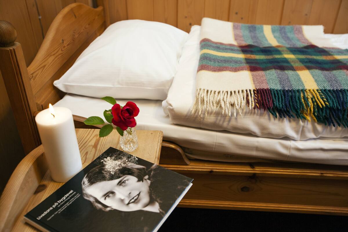 Una cama de madera con un almohadón blanco y una manta colorida. Una vela blanca y un libro están sobre una mesita de noche de madera al lado de la cama.
