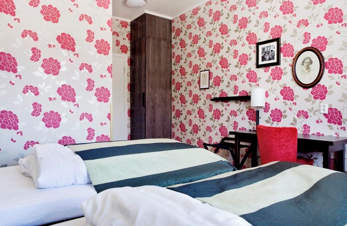 Una habitación con papel tapiz floral, una cama con ropa de cama a rayas, un escritorio con una silla y un cuadro en la pared.
