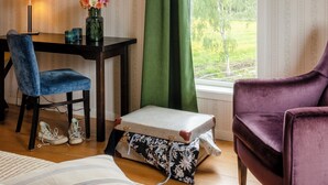 Una habitación con una cama, un escritorio, una silla, un sillón morado, una maleta y una ventana con vistas a la vegetación.