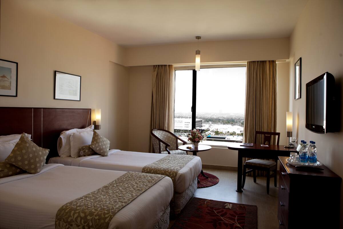 Habitación de hotel con dos camas, un escritorio, una silla, televisor y ventana con cortinas.