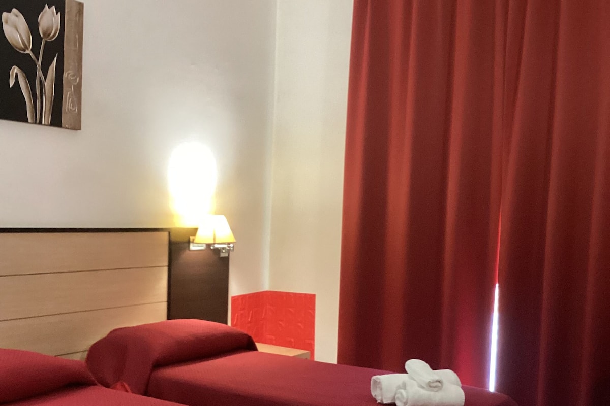 Ein Hotelzimmer mit zwei Betten, roter Bettwäsche, einem Nachttisch mit Lampe, einem Bild an der Wand und einem roten Vorhang.