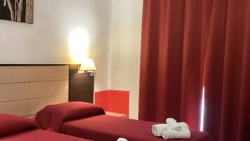 Habitación de hotel con dos camas, ropa de cama roja, una mesita de noche con lámpara, un cuadro en la pared y una cortina roja.