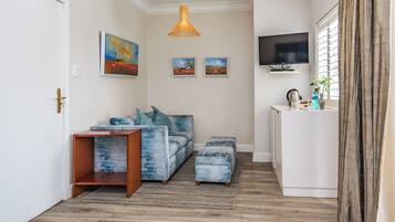 Una sala de estar moderna con un sofá, un reposapiés y una pequeña mesa de madera. Hay tres cuadros enmarcados en la pared, un televisor y una ventana con cortinas.