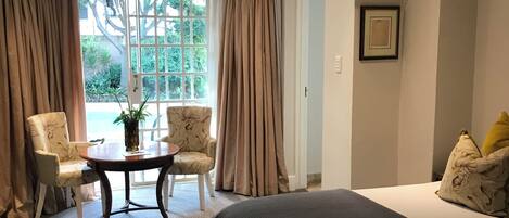 A bedroom with a large bed, two chairs, a small table, and a view of greenery outside.