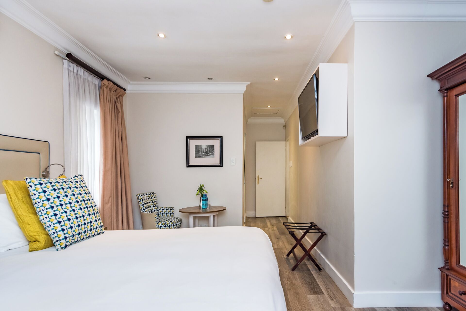 A hotel room with a bed, a small table, a chair, a TV mounted on the wall, and a wardrobe.