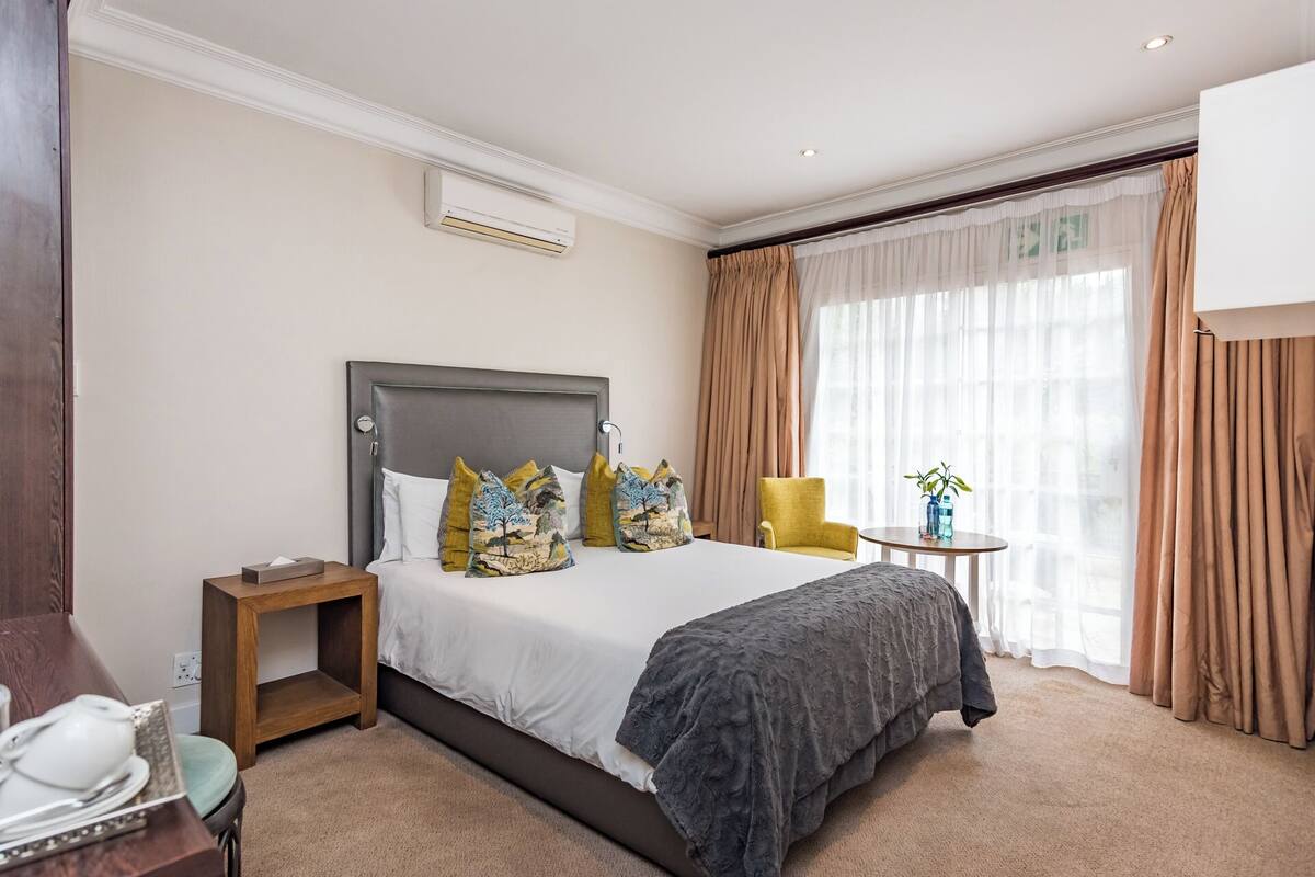A hotel room with a large bed, a bedside table, a chair, a small table with a vase, and a window with curtains.