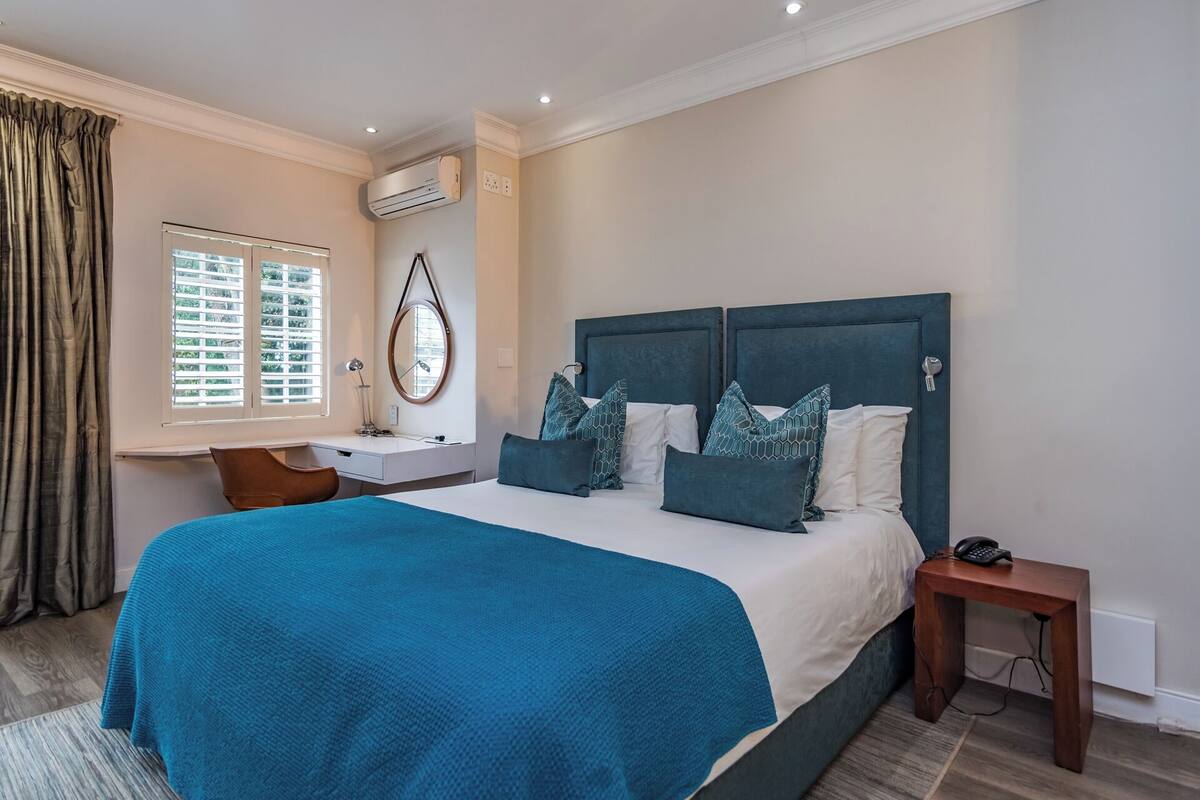 A bedroom with a large bed, a bedside table, a desk, a chair, an air conditioning unit, and a window with blinds.