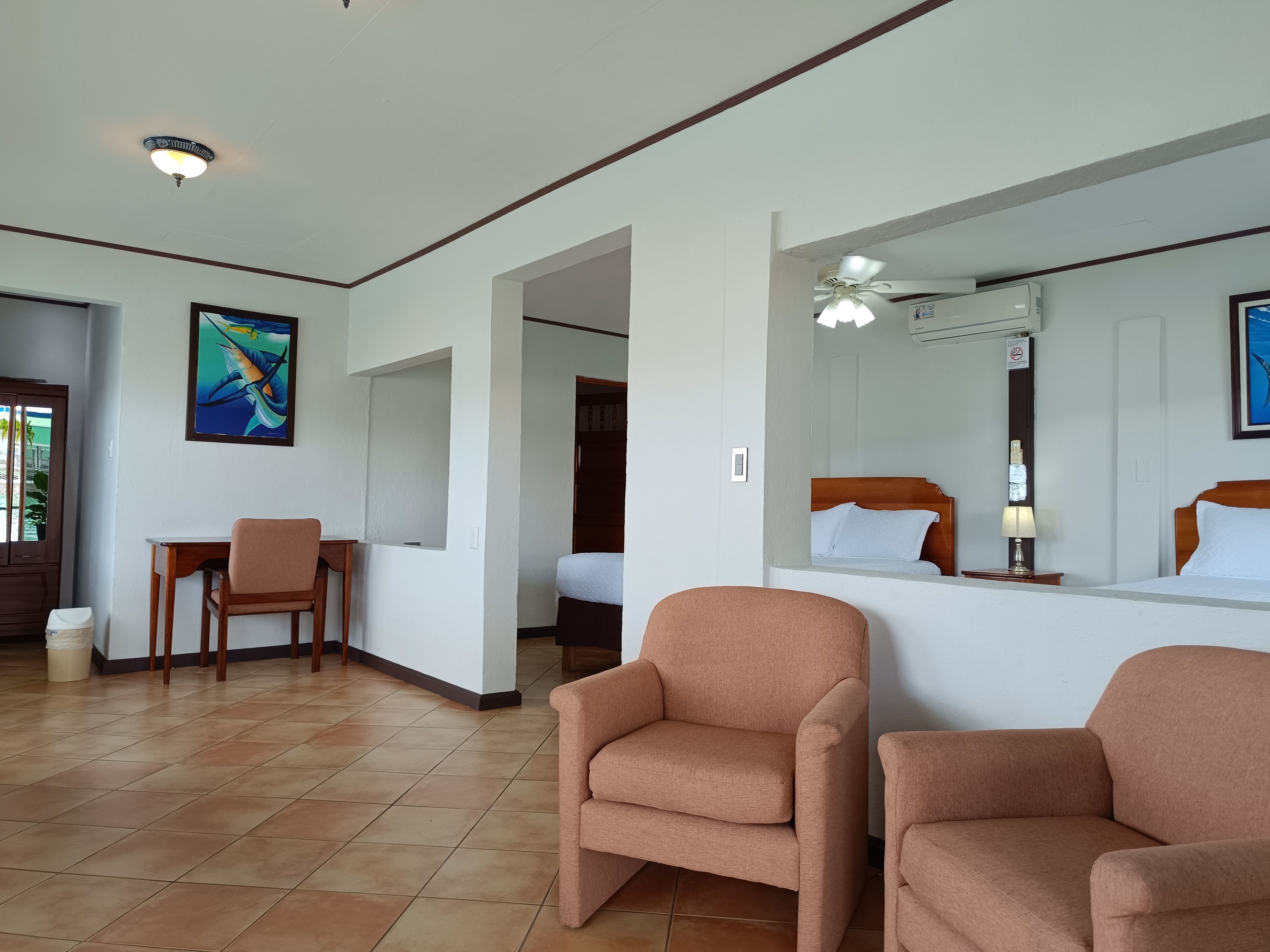 Une chambre d’hôtel comprenant un lit, deux fauteuils, un bureau, une chaise, un tableau et un ventilateur de plafond.