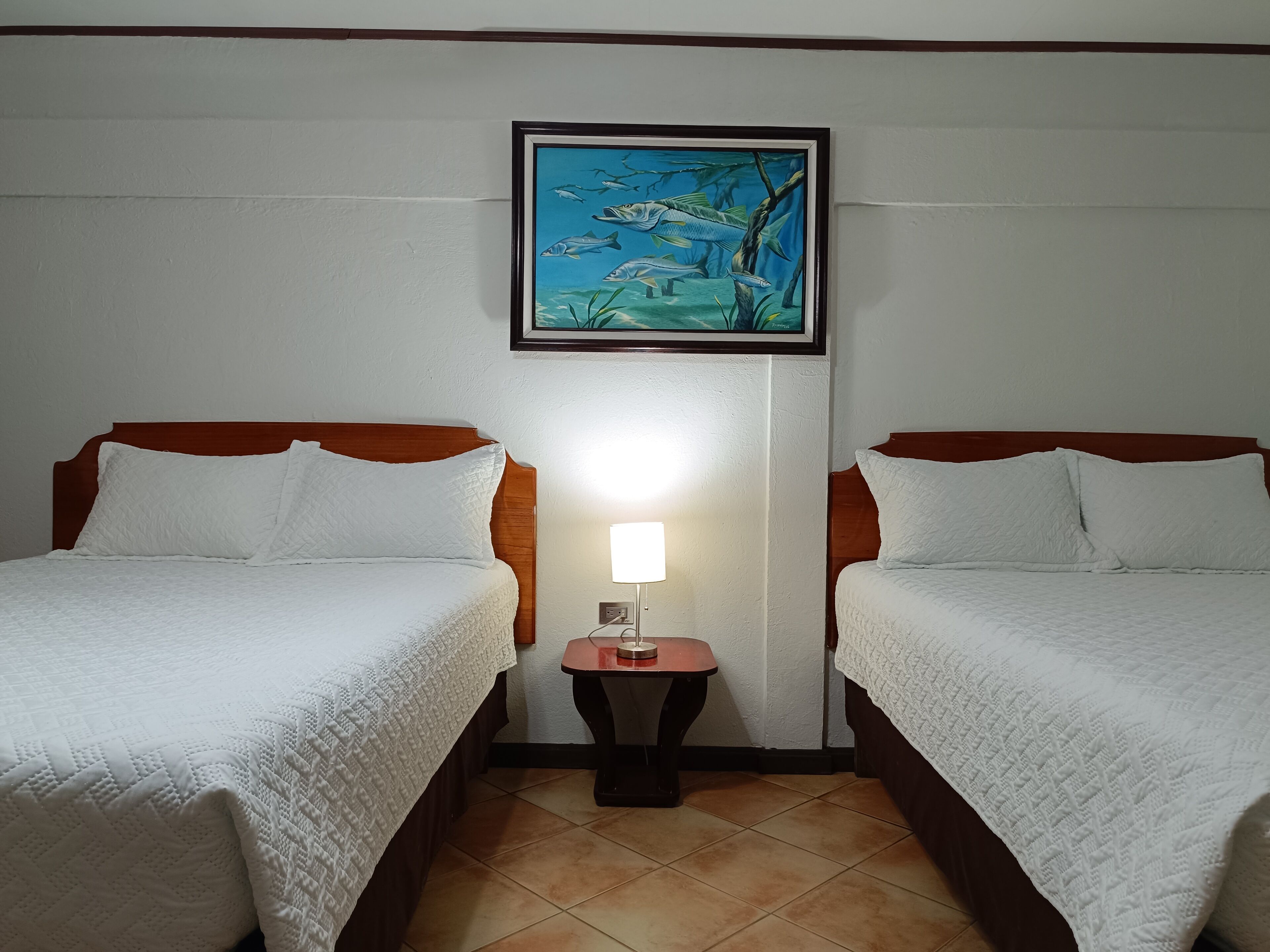 A hotel room with two beds, a nightstand with a lamp, and a framed picture of fish on the wall.
