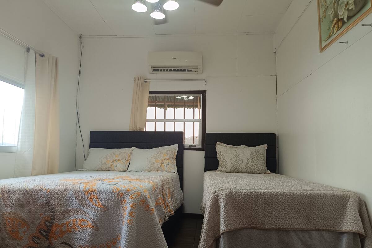 A room with two beds, a window with curtains, an air conditioning unit, and a ceiling light fixture.