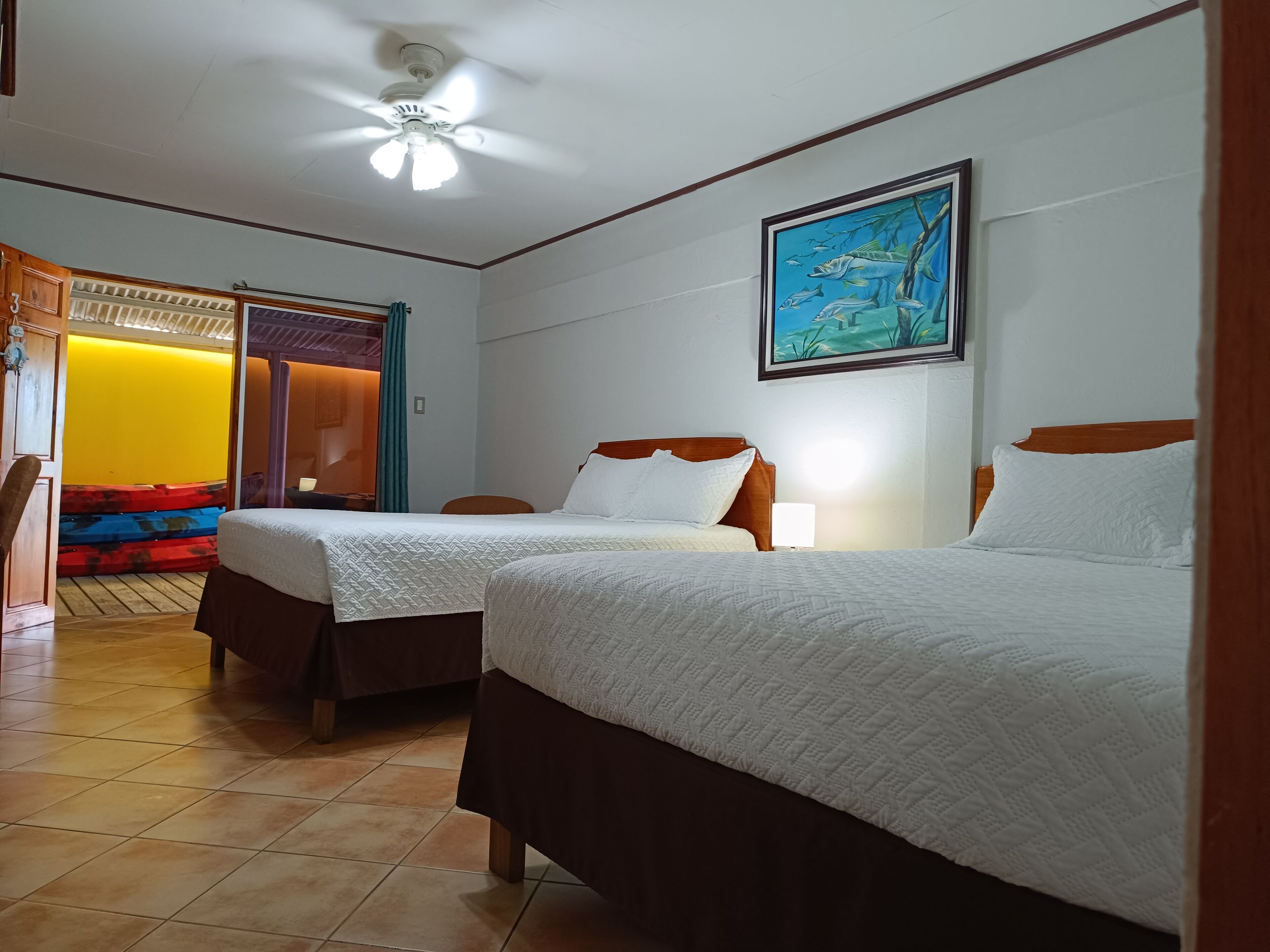 Quarto de hotel com duas camas, um ventilador de teto, um quadro e vista para o exterior.