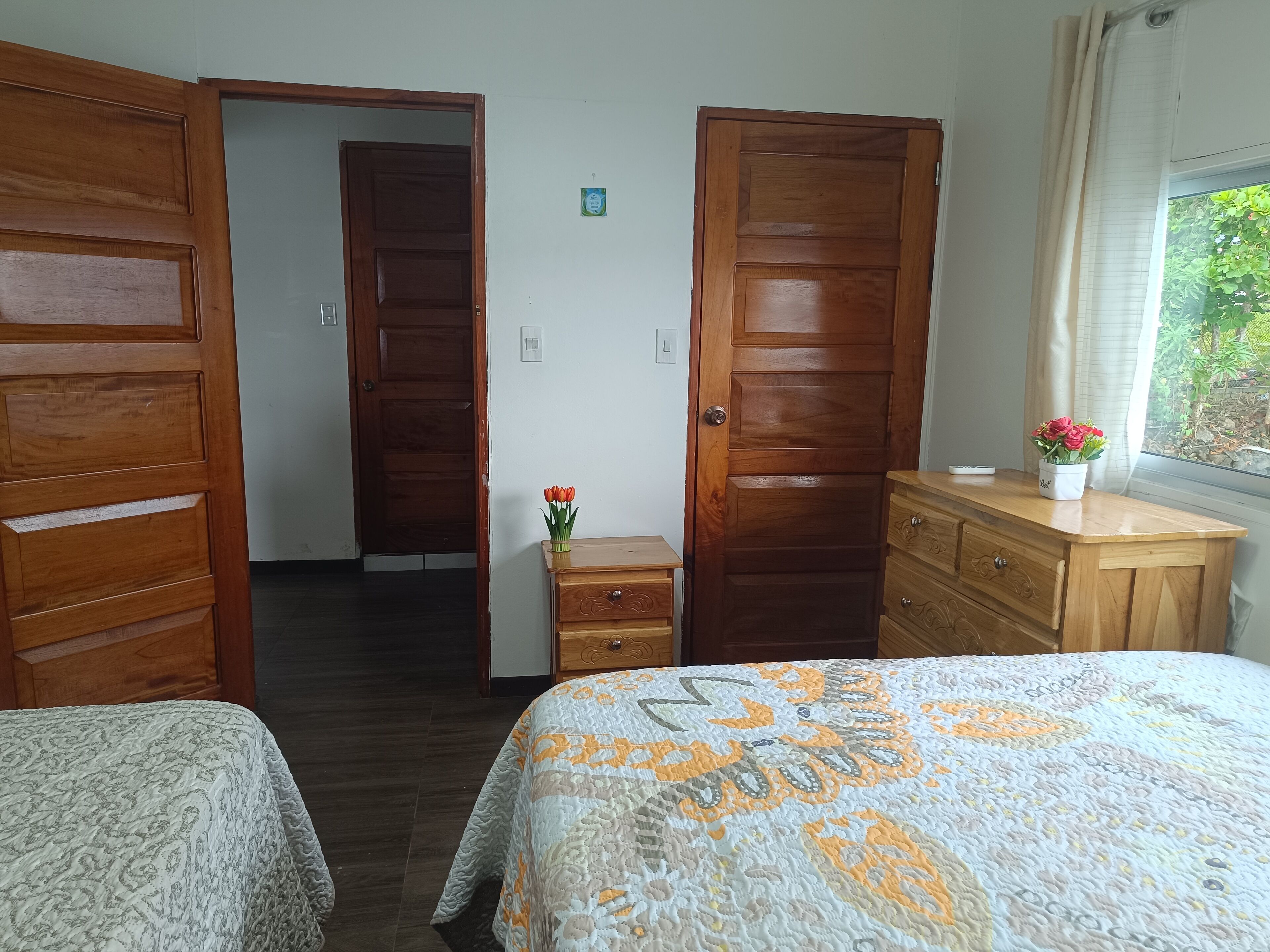 A hotel room with two wooden doors, a wooden dresser, a bedside table with a vase of flowers, and a patterned bedspread.
