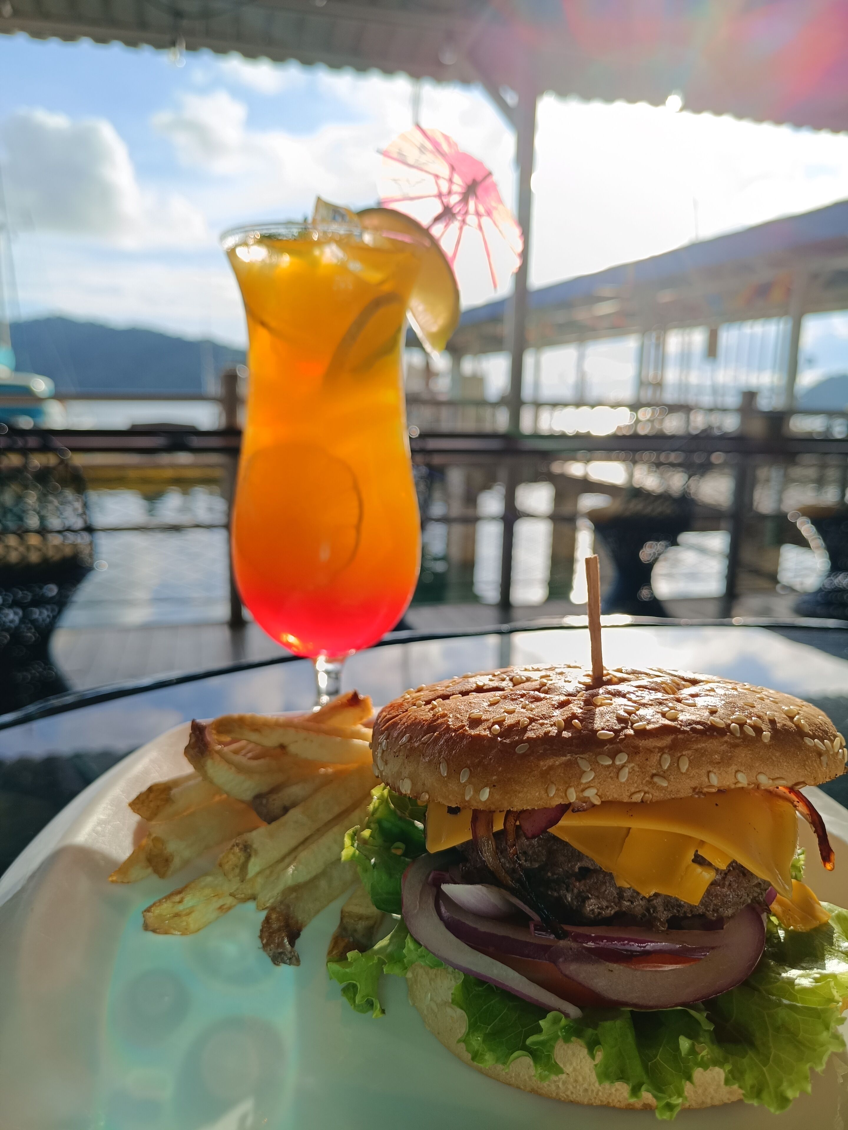A burger with a sesame seed bun, lettuce, red onion, cheese, and a beef patty, served with fries and a glass of orange drink with a straw and a slice of lime.