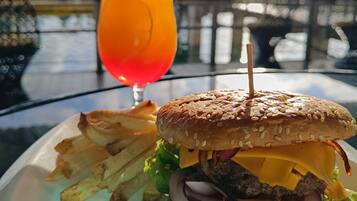 A burger with a sesame seed bun, lettuce, red onion, cheese, and a beef patty, served with fries and a glass of orange drink with a straw and a slice of lime.