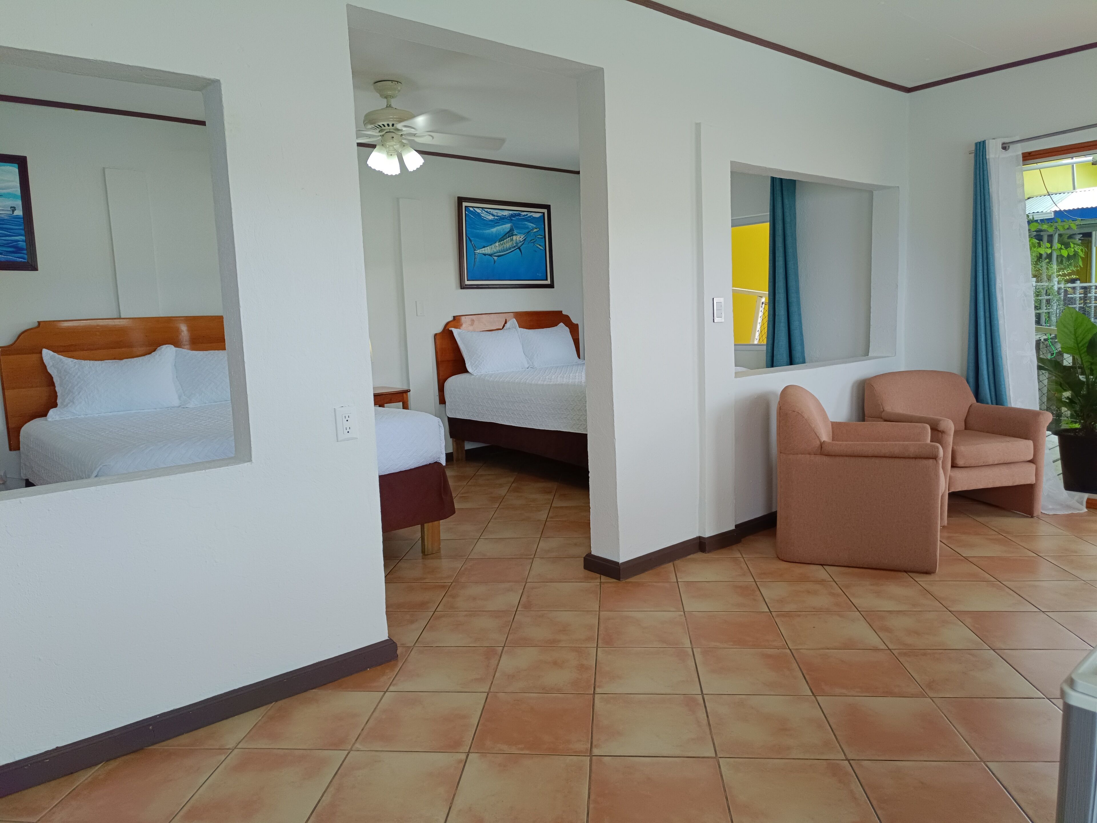 Habitación de hotel con dos camas, ventilador de techo, un espejo grande y un área de descanso con una silla y una mesa.