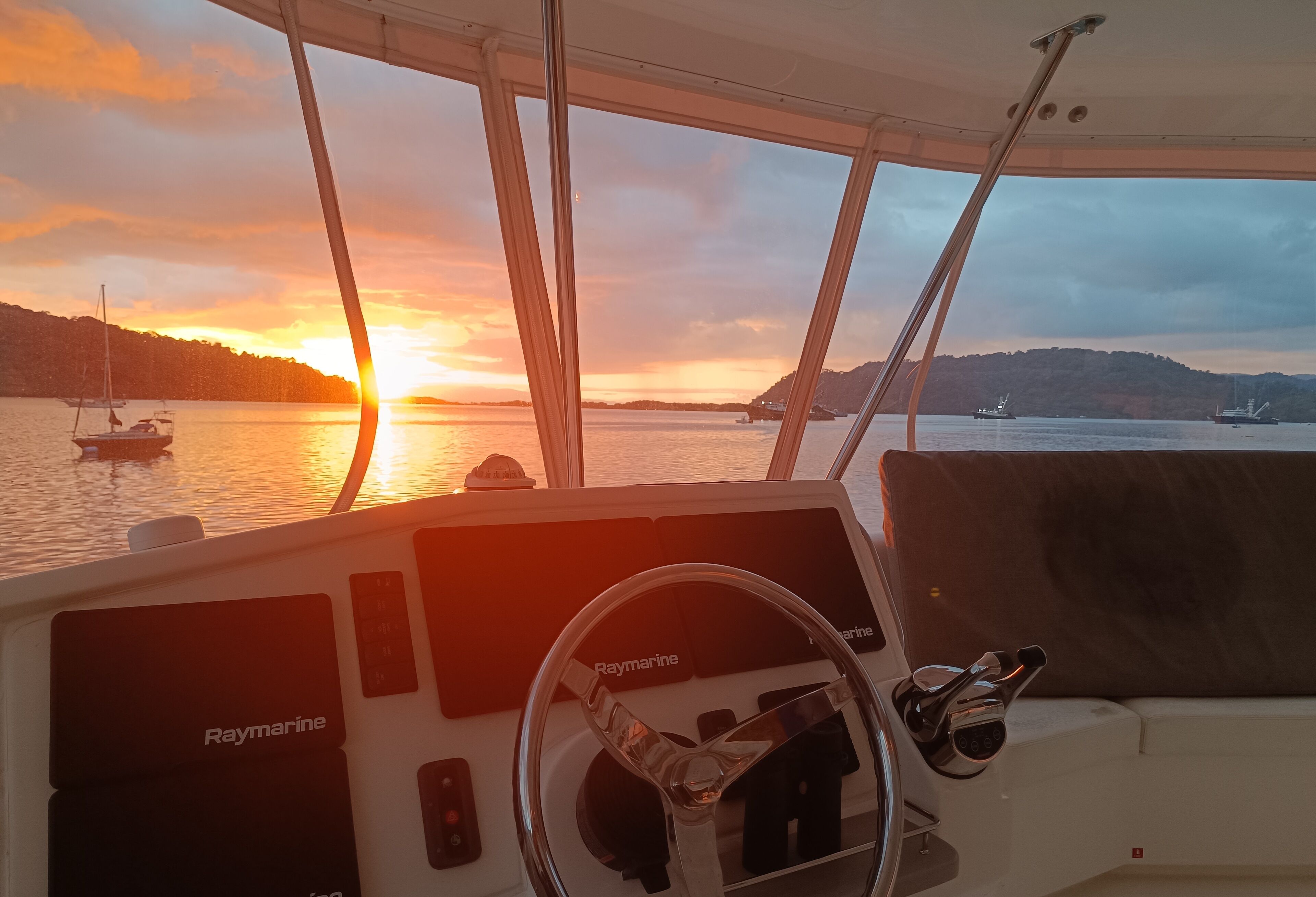 A boat's interior with a steering wheel labeled "Raymarine," a control panel, and a view of the sunset over water with boats and hills in the background.