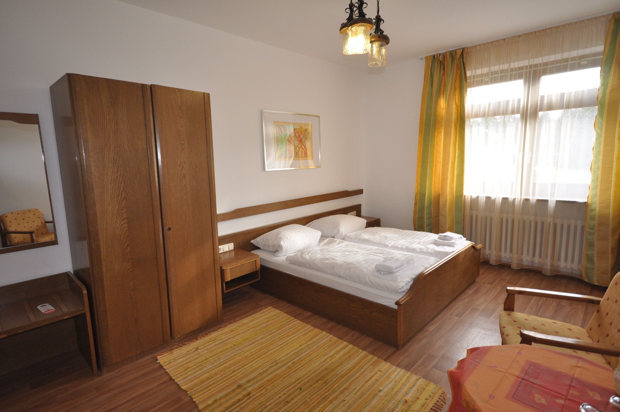Ein Schlafzimmer mit einem hölzernen Bett, einem Nachttisch, einem Kleiderschrank, einem Sessel und einem Fenster mit Vorhängen.