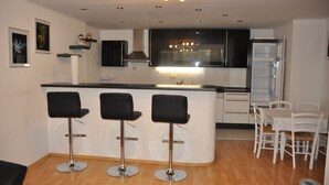 Een moderne keuken met een centraal kookeiland, drie zwarte barkrukken, een eettafel met vier stoelen en een koelkast.