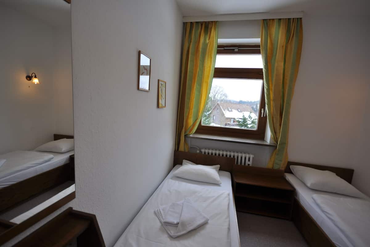 Una habitación pequeña y limpia con dos camas individuales, una ventana con cortinas, un radiador y una lámpara fijada a la pared.