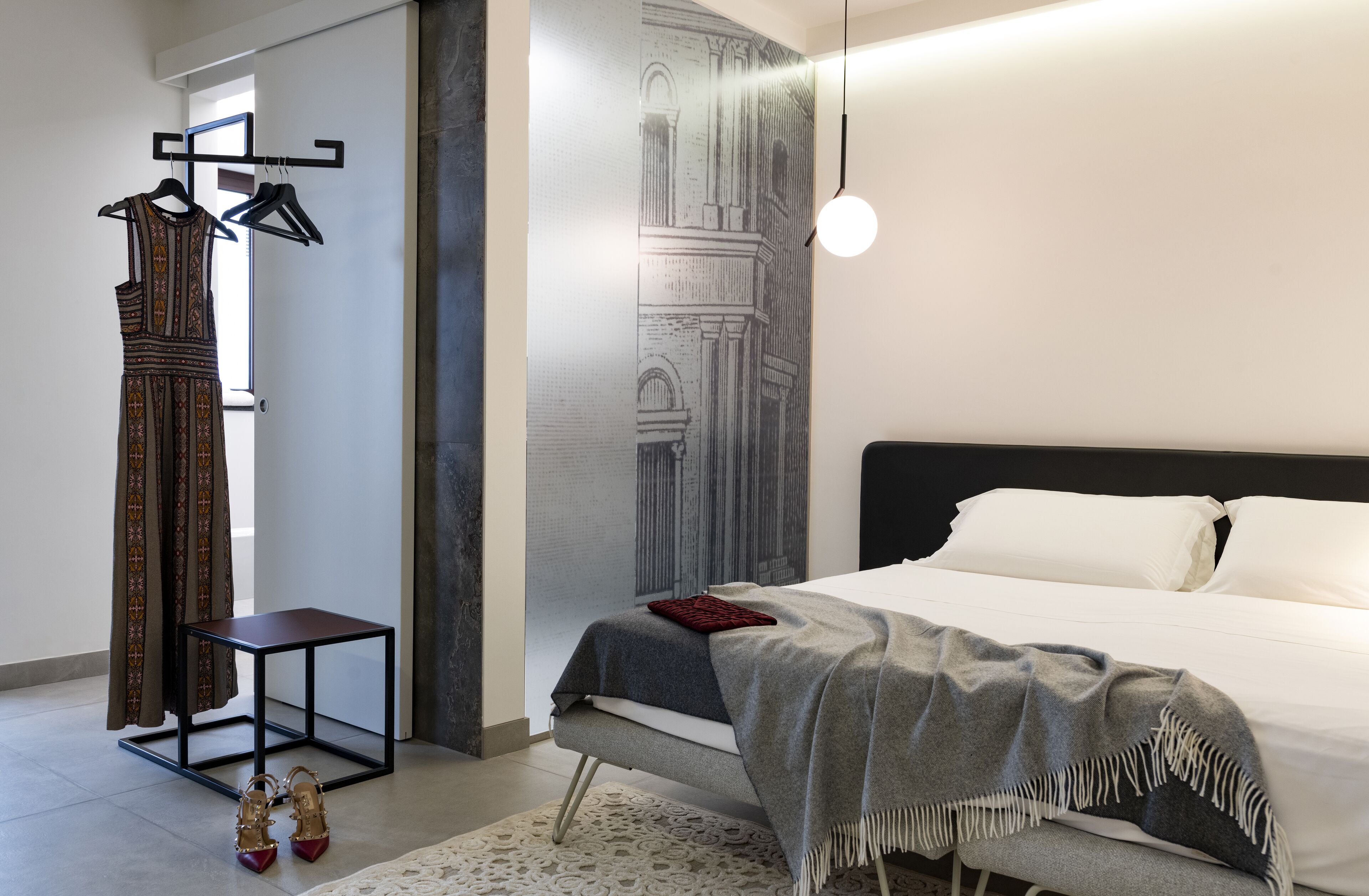 Un dormitorio moderno con cama, mesita de noche, luz de pared y un vestido colgado.