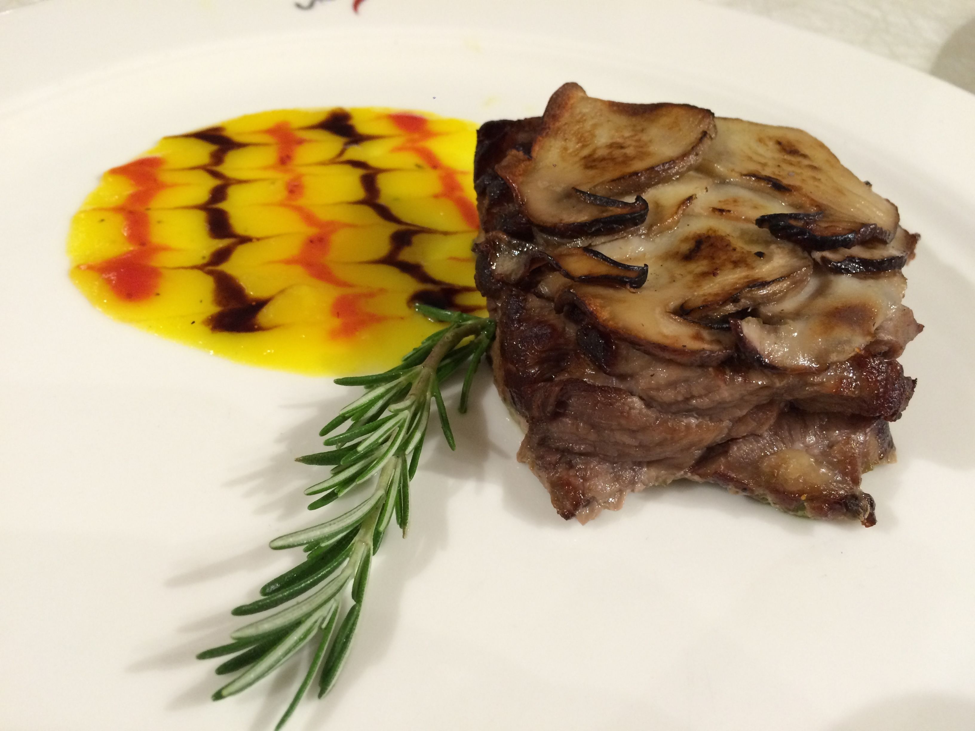 A plate with a piece of meat topped with mushrooms and a yellow sauce with a red pattern.