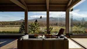 Una sala de estar moderna con sofá de madera, una mesa de comedor y una silla. Cuenta con ventanas amplias que ofrecen vistas a una cadena montañosa nevada.