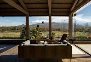Una sala de estar moderna con sofá de madera, una mesa de comedor y una silla. Cuenta con ventanas amplias que ofrecen vistas a una cadena montañosa nevada.