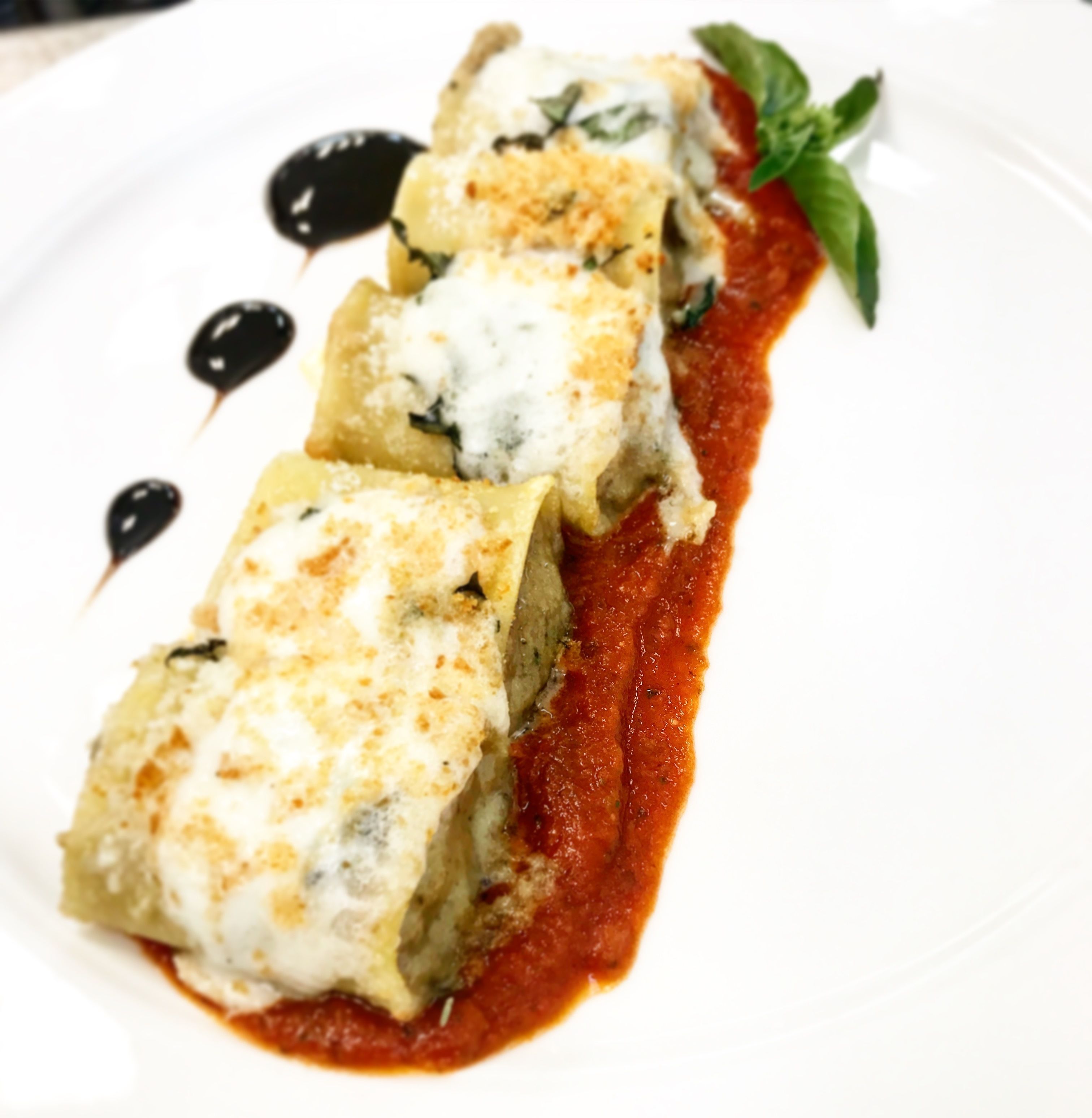 A plate with three stuffed pasta rolls topped with cheese and a basil leaf, served with a red sauce and black olive garnish.