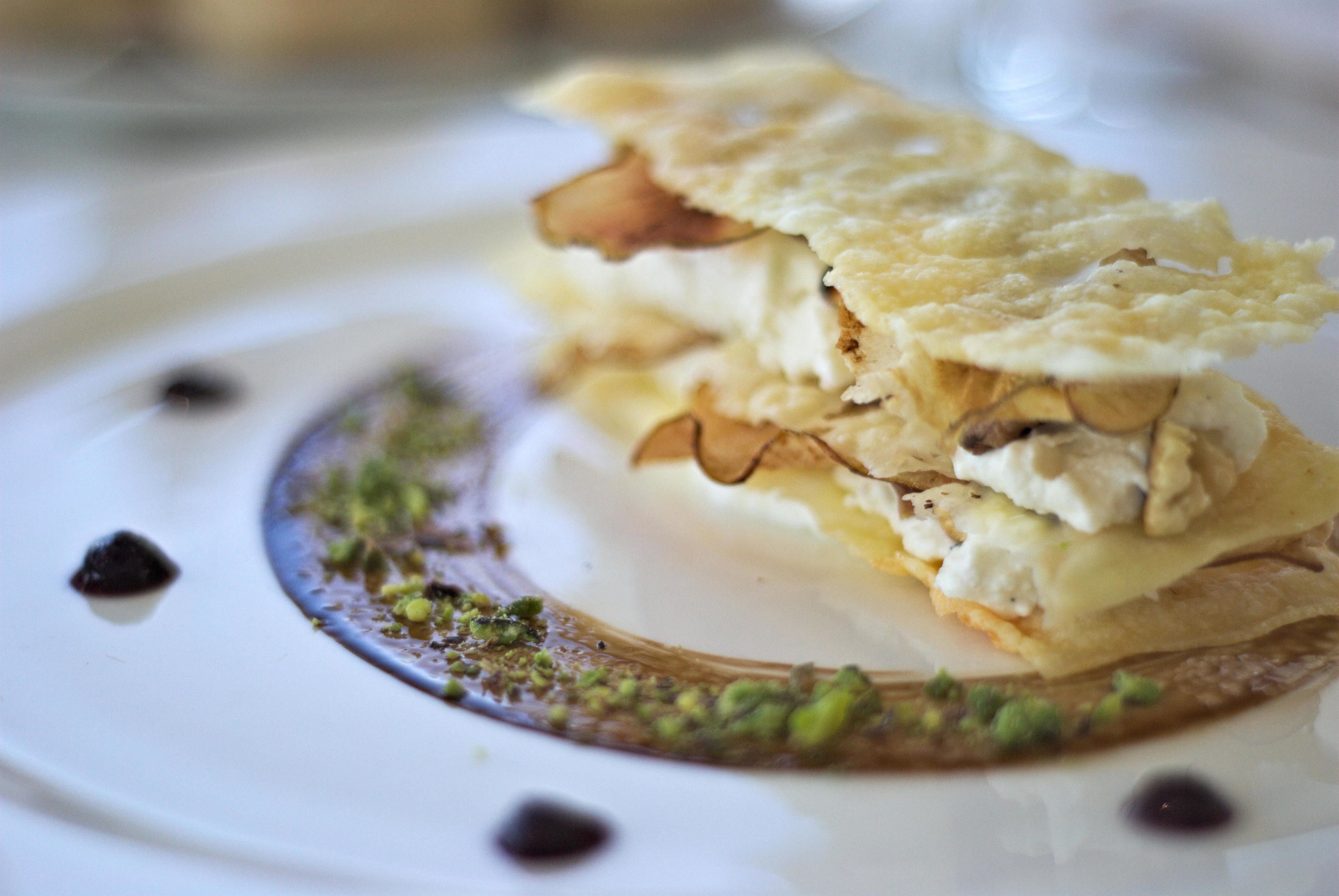 A dessert with a pastry base, topped with whipped cream and nuts, on a white plate with a decorative sauce and green garnish.